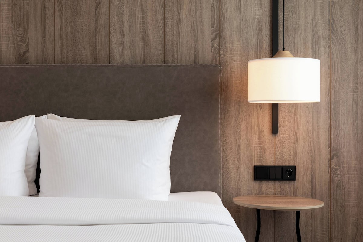 Una cama bien hecha con sábanas y almohadas blancas, una mesita de madera redonda y una lámpara fijada a la pared.