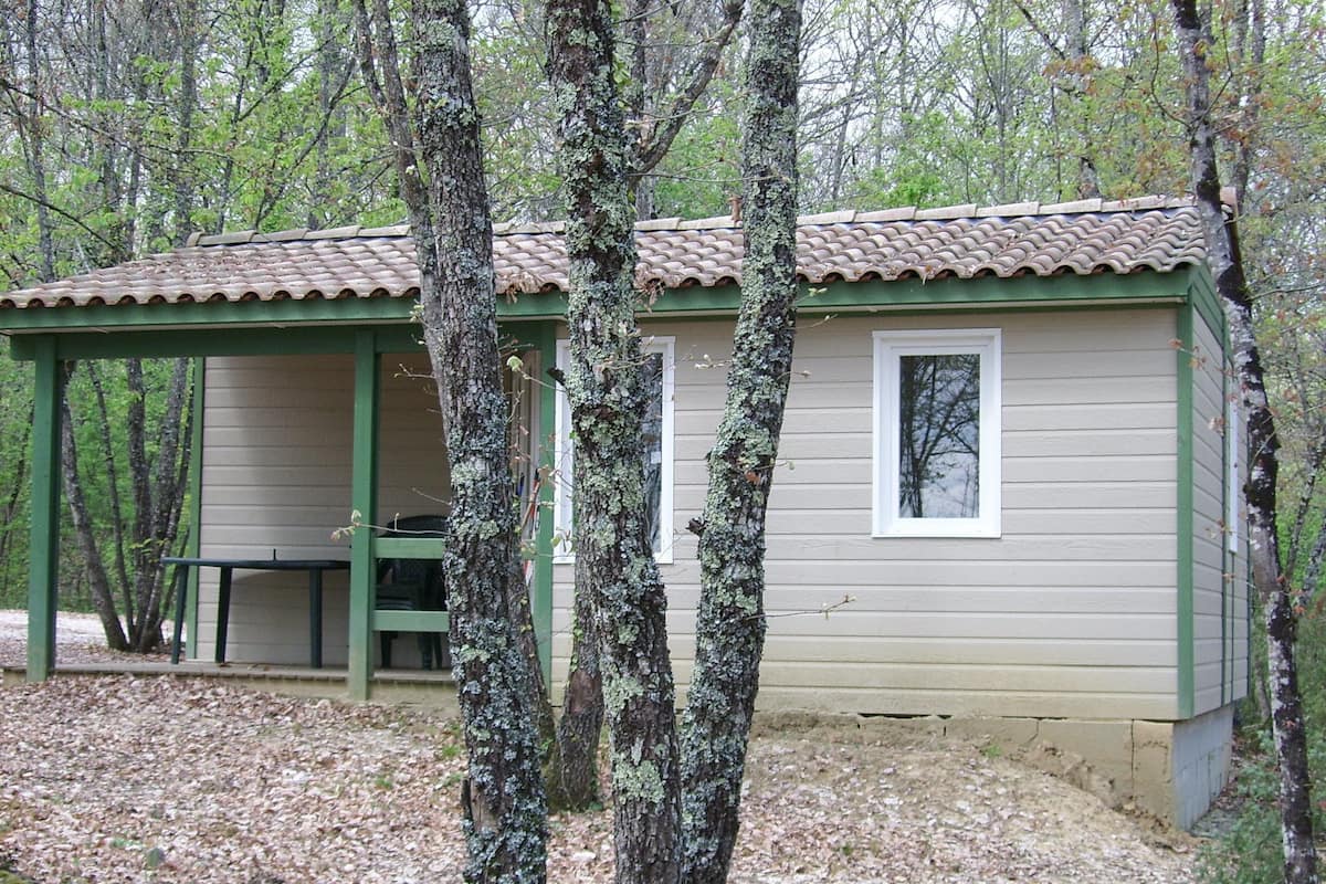 Une petite cabane avec un toit vert et une fenêtre, entourée d’arbres et dotée d’un espace abrité avec un banc.