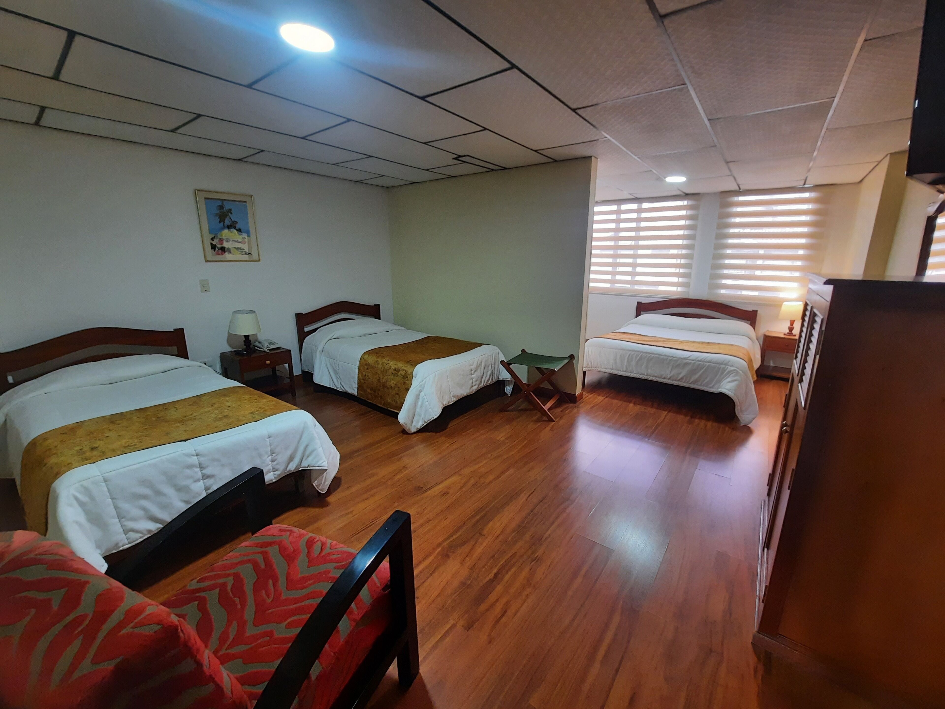 A hotel room with three beds, wooden flooring, and a ceiling with recessed lighting.