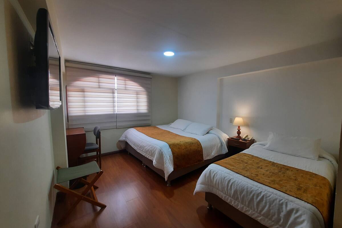 A hotel room with two beds, a desk, a chair, a TV, and a window with blinds.
