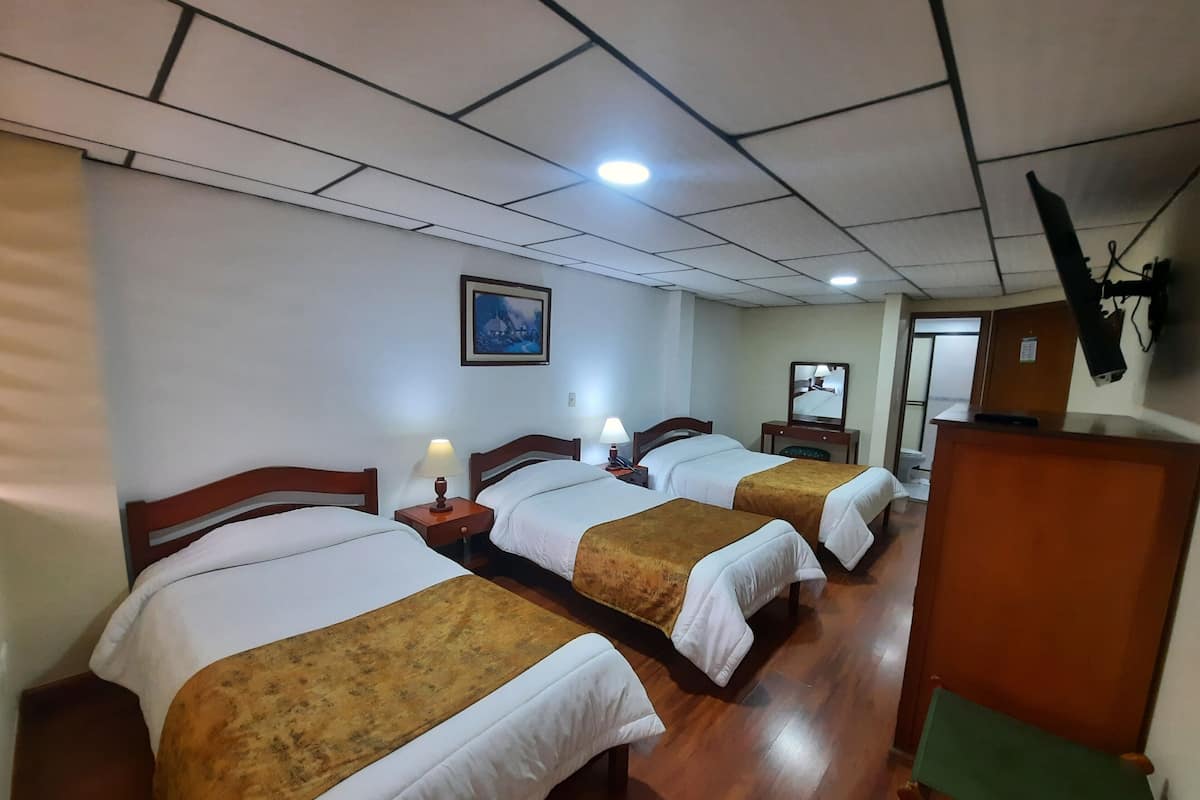 A hotel room with three beds, wooden headboards, a painting on the wall, and a television mounted on the wall.