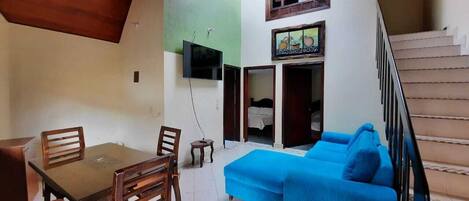 A room with a dining table and chairs, a blue sofa, a TV mounted on the wall, and a staircase.