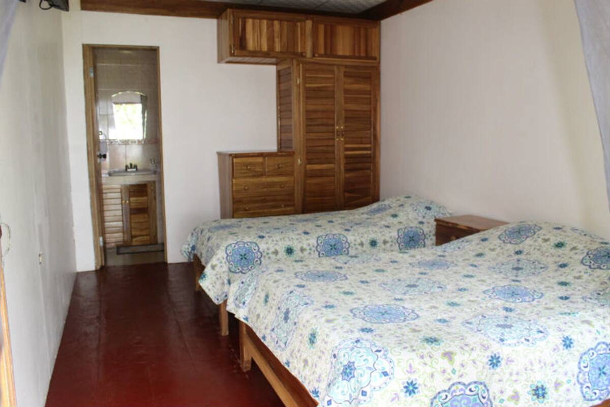 A bedroom with a wooden wardrobe, a bed with patterned bedding, and a small bathroom with a mirror and sink.