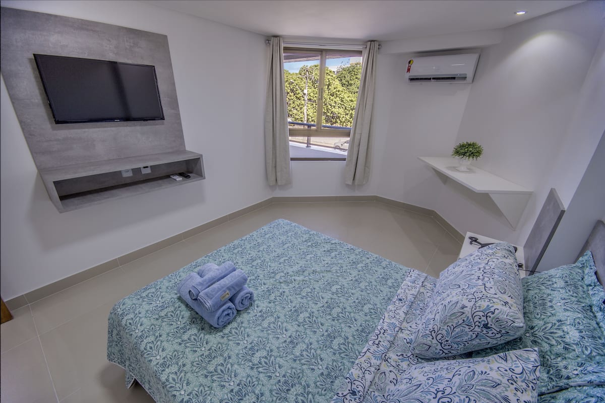 A bedroom with a bed, a flat-screen TV mounted on the wall, a window with curtains, a small shelf with a plant, and an air conditioning unit.