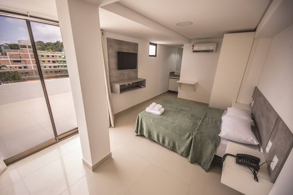 A modern hotel room with a large window, a bed with a green bedspread, a bedside table with a phone, and a built-in TV.