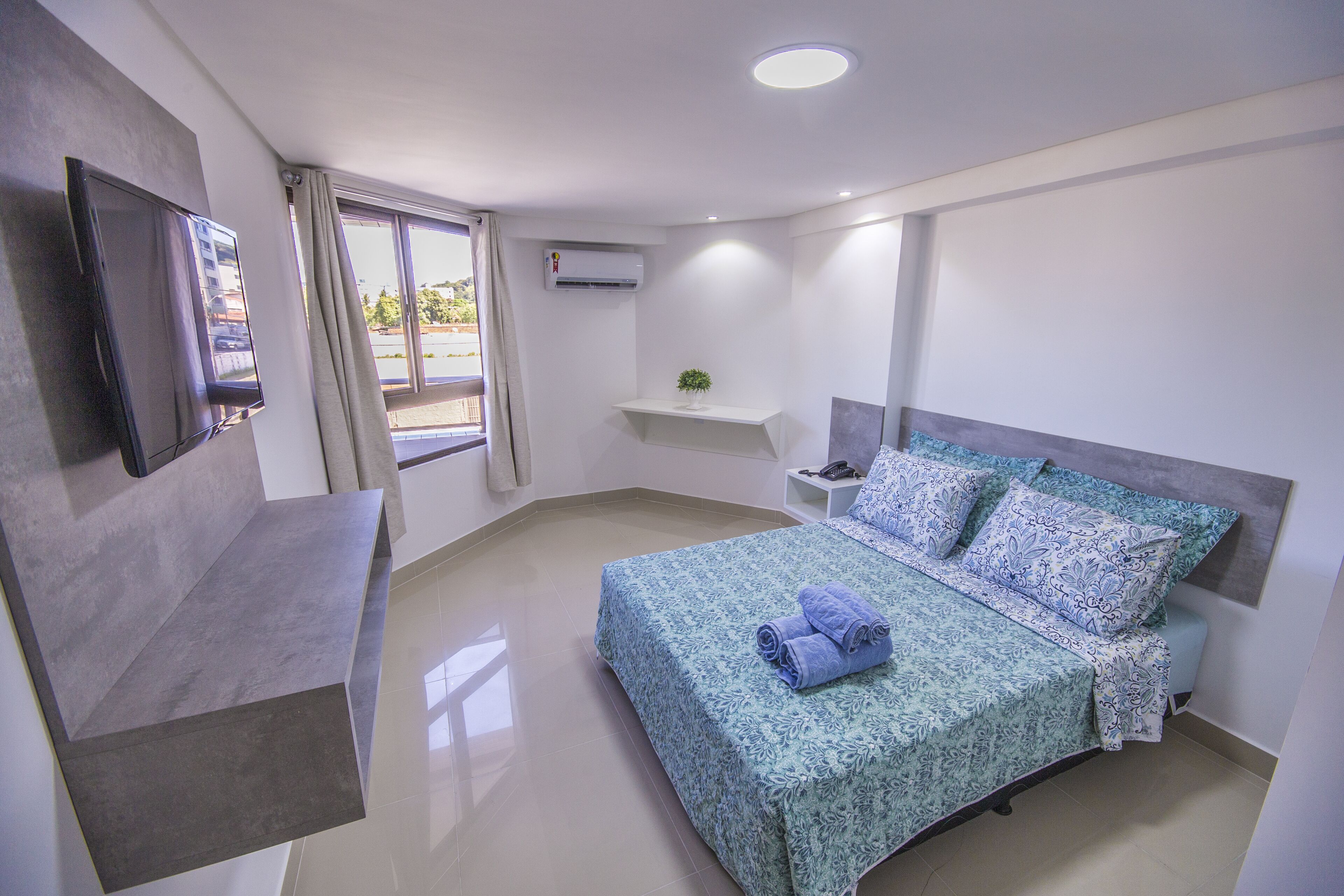 Una habitación de hotel moderna con una cama grande, un televisor de pantalla plana montado en la pared, una mesita auxiliar y una ventana con cortinas.