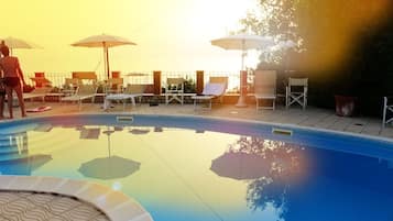 Una piscina con lettini e ombrelloni, circondata da un muro di pietra e un recinto metallico.