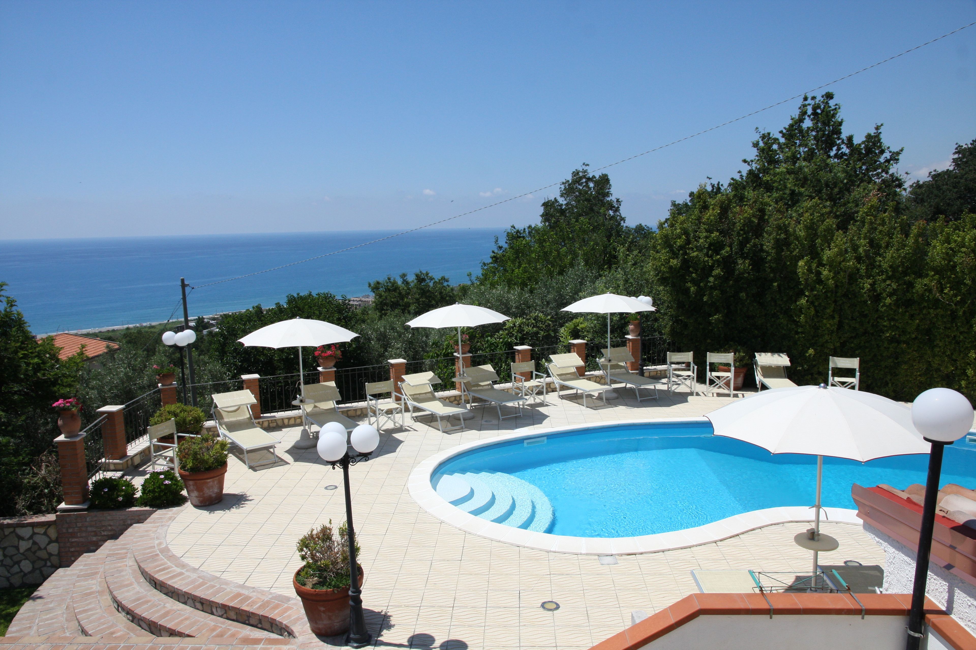 Una piscina con sillas de descanso y sombrillas, rodeada de plantas en macetas y vistas al mar.