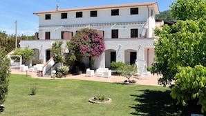 A two-story building with a white exterior and a red roof, featuring a patio area with white furniture and a large flowering tree. - Villa Catalano (Paola)