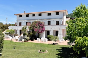 A two-story building with a white exterior and a red roof, featuring a patio area with white furniture and a large flowering tree. - Villa Catalano (Paola)