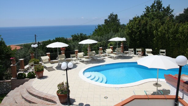 Ein Swimmingpool mit Liegestühlen und Sonnenschirmen, umgeben von Topfpflanzen und Blick auf das Meer.