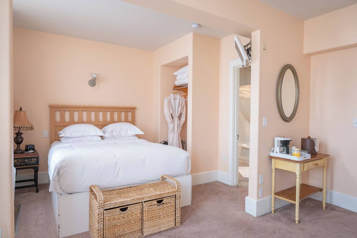 A bedroom with a bed, a bedside table with a lamp, a small table with a coffee maker, and a wicker basket.