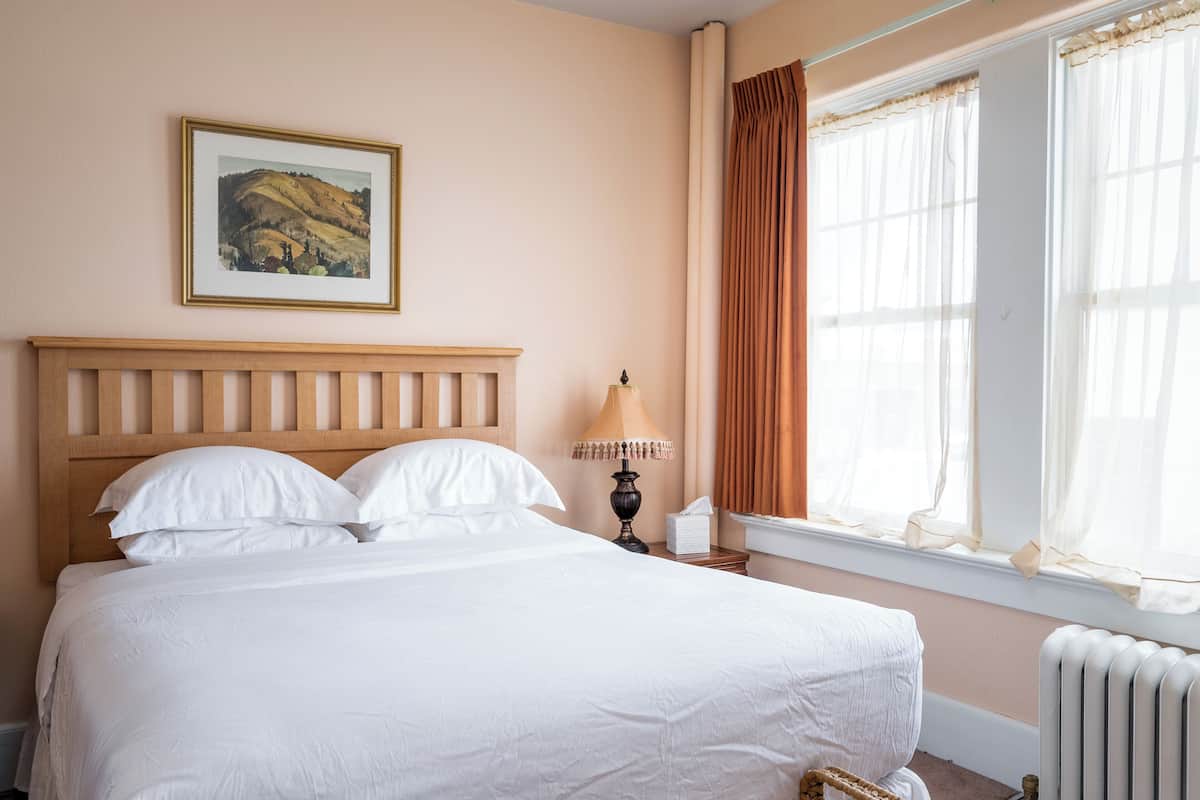 A neatly made bed with white linens in a room with a wooden headboard, a framed landscape painting on the wall, and a window with sheer curtains.