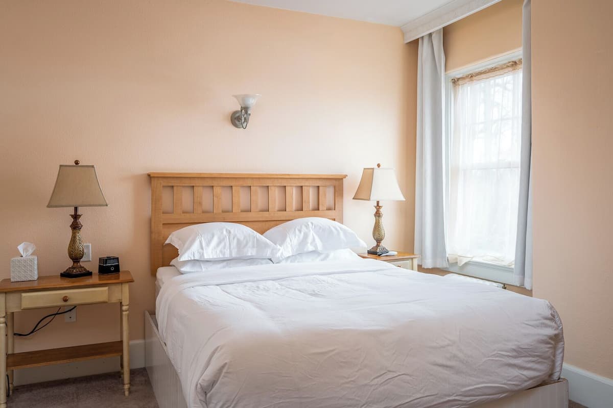 A neatly made bed in a room with peach walls, a wooden headboard, two bedside tables with lamps, a nightstand, and a window with sheer curtains.