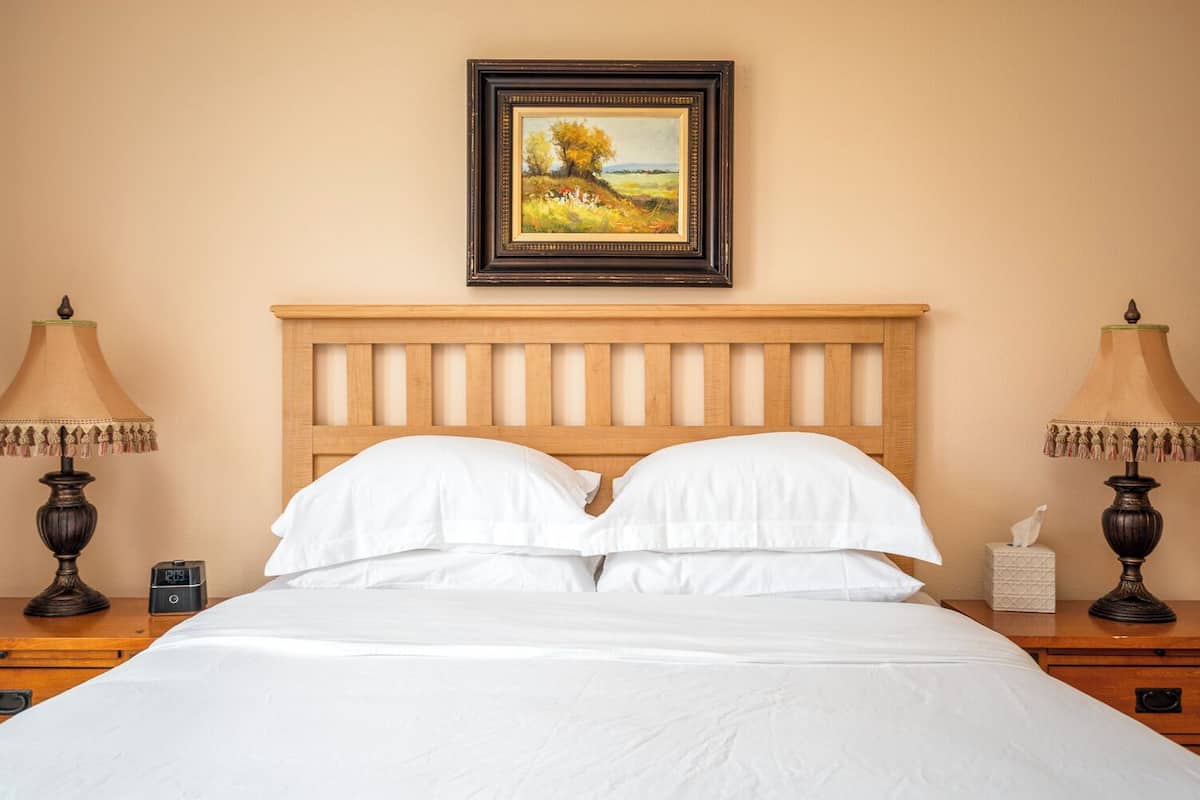 A neatly made bed with white linens, a wooden headboard, and a framed landscape painting above it.