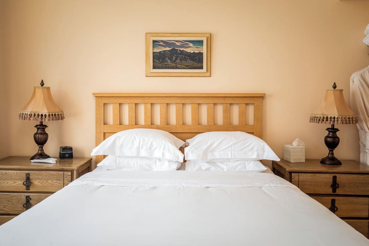 A neatly made bed with a wooden headboard, two bedside tables with lamps, a small nightstand, and a framed picture on the wall.