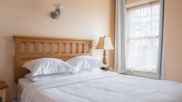 A neatly made bed with white linens in a room with a wooden headboard, a wall-mounted light fixture, a bedside table with a lamp, and a window with sheer curtains.