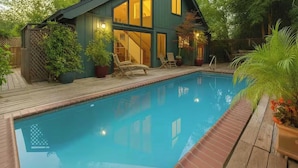 Une piscine avec une terrasse en bois, une maison dotée de grandes fenêtres et des plantes en pot.