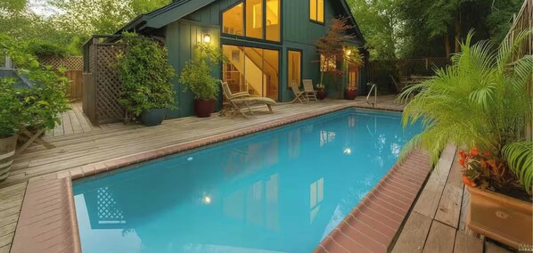 Una piscina con muelle de madera, una casa con ventanales grandes y plantas en macetas.