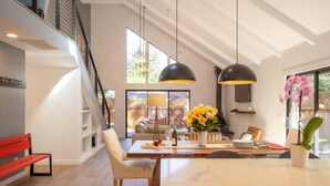 Une cuisine moderne avec un coin repas, comprenant une table en bois, des chaises, des luminaires suspendus et une vue sur l’extérieur.