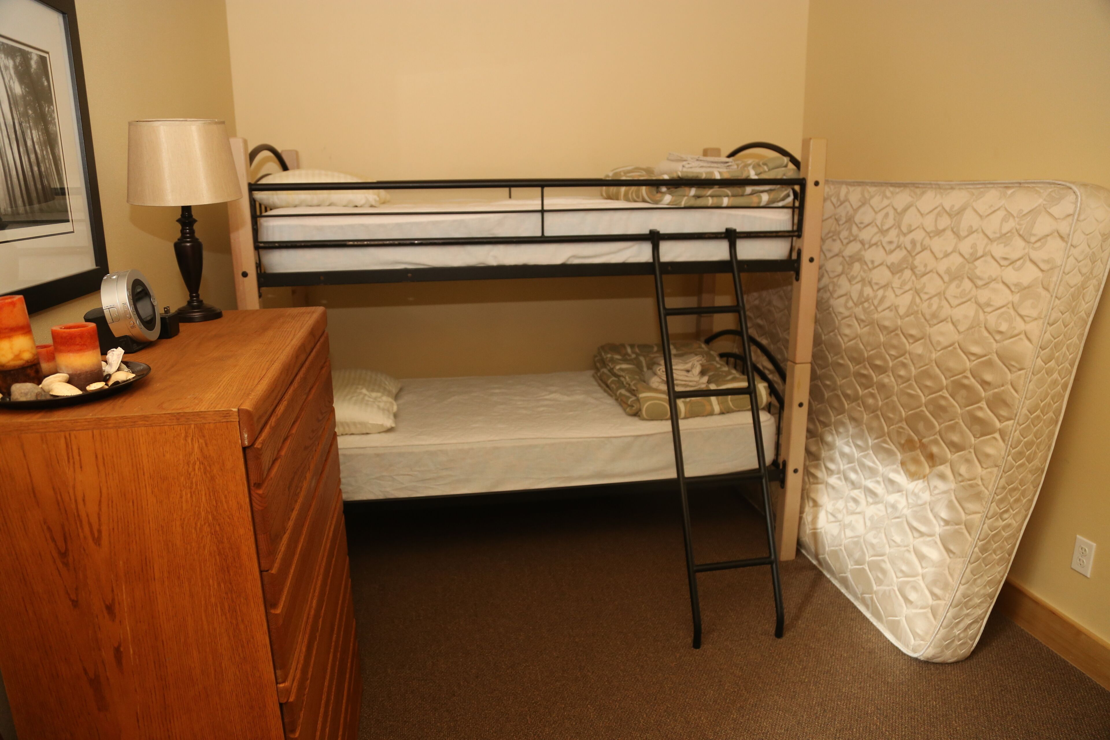 A bunk bed with a mattress on the floor, a wooden dresser, a lamp, and a framed picture on the wall.