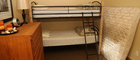 A bunk bed with a mattress on the floor, a wooden dresser, a lamp, and a framed picture on the wall.