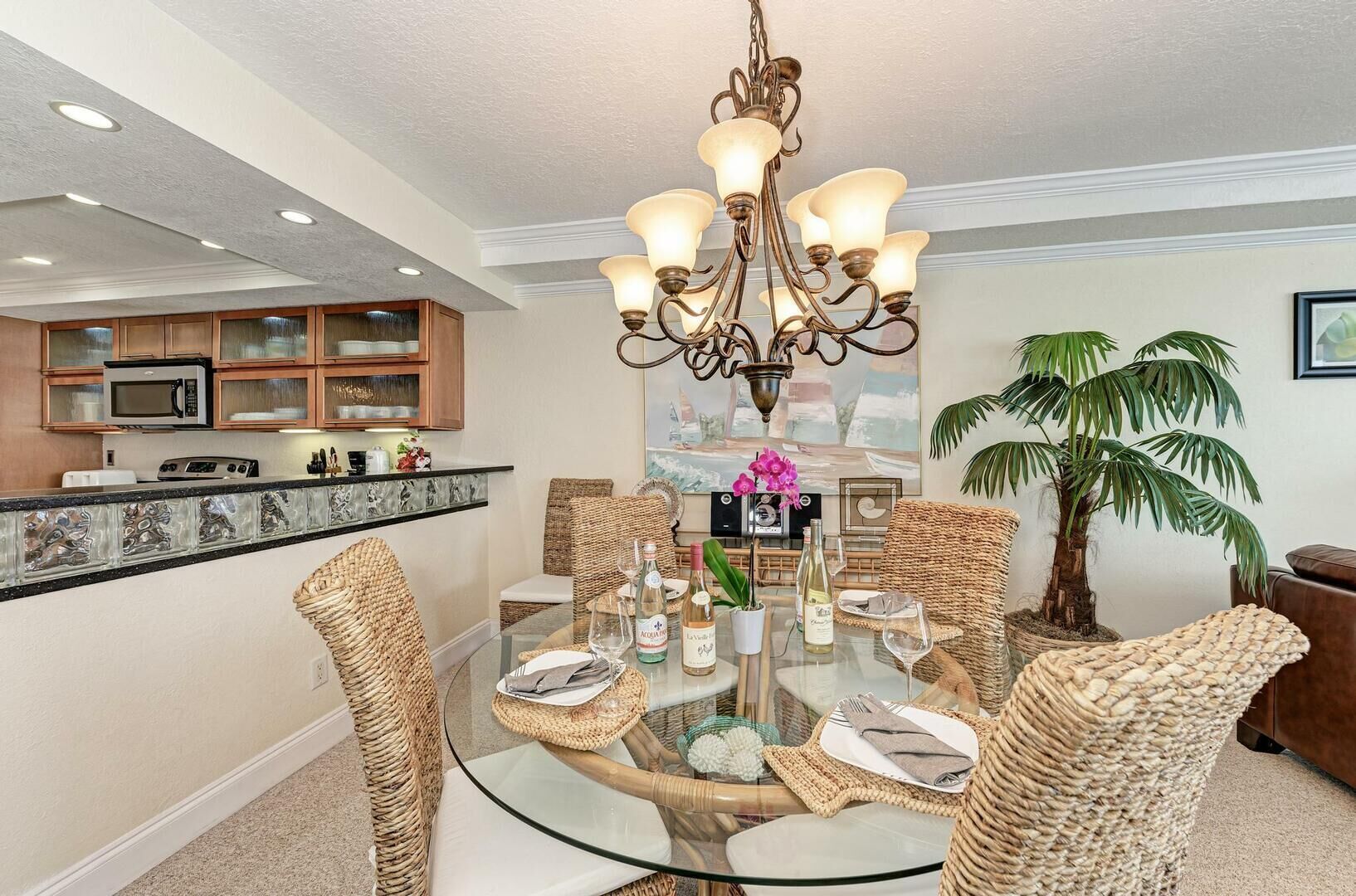 A dining area with a glass table surrounded by wicker chairs, a chandelier, and a kitchen in the background.