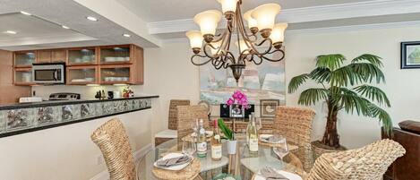 A dining area with a glass table surrounded by wicker chairs, a chandelier, and a kitchen in the background.