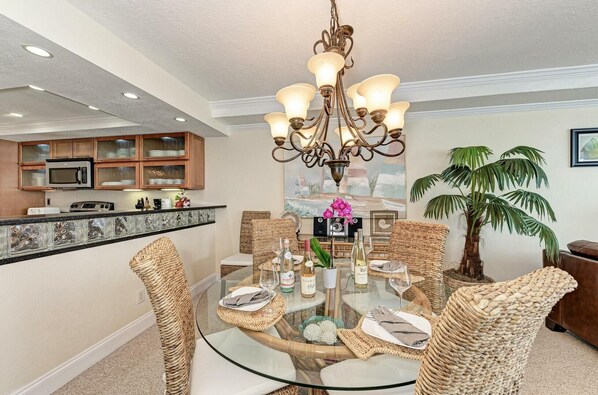 A dining area with a glass table surrounded by wicker chairs, a chandelier, and a kitchen in the background.
