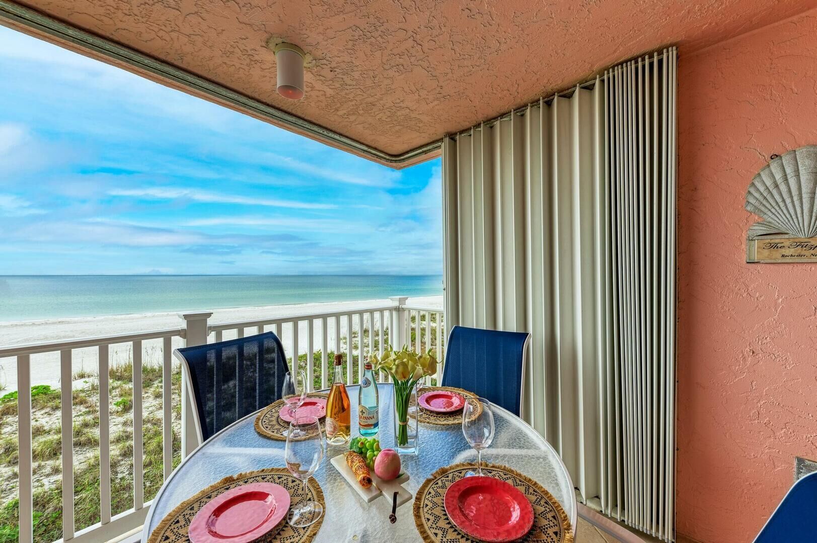 Ein runder Tisch mit Glasplatte, gedeckt für zwei Personen, umgeben von weißen Stühlen mit Blick auf einen Strand und den weiten Ozean.