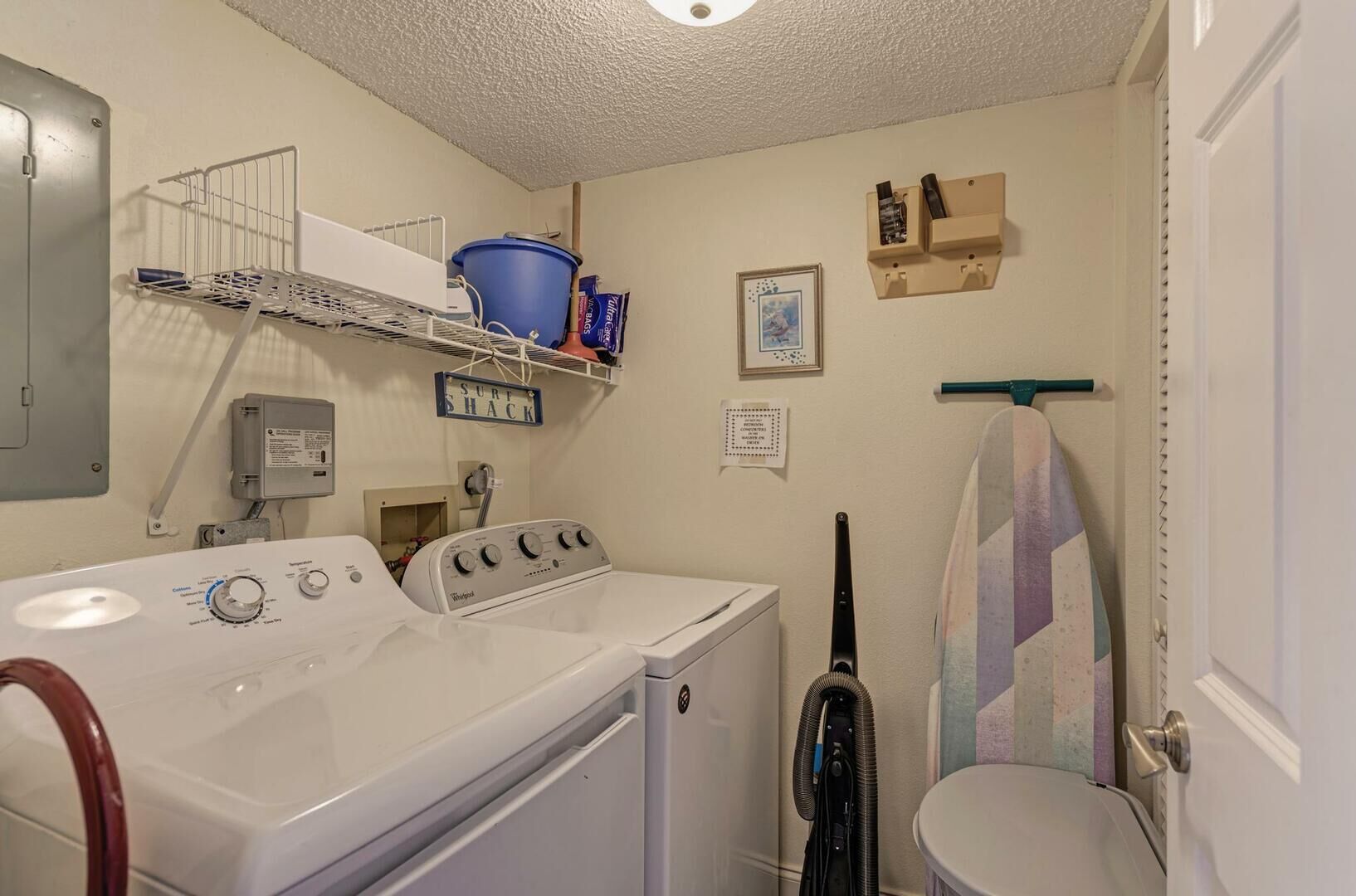 A laundry room with a front-loading washer and dryer, a shelf with laundry supplies, a blue bucket, and a wall-mounted heater.