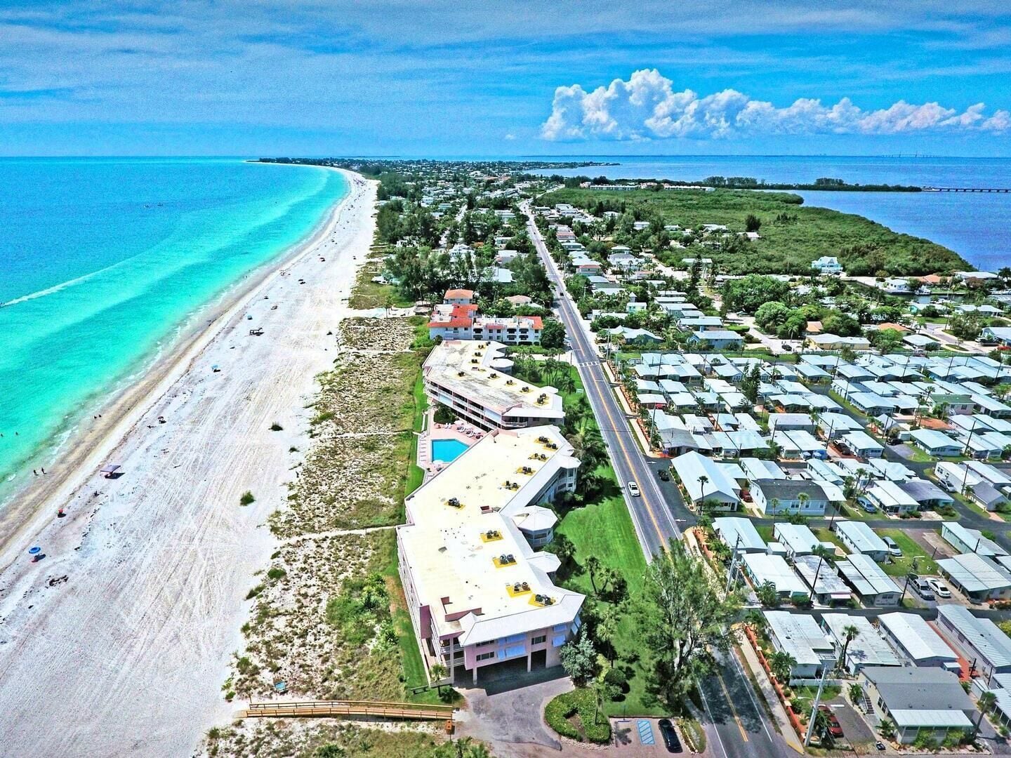An aerial view of a coastal area with a beach, a row of mobile homes, and a large building with a swimming pool.