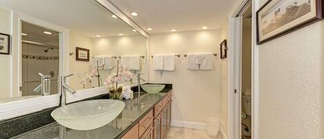 A modern bathroom with a large glass sink, two mirrors, a vanity with drawers, and a shower area visible through a glass door.