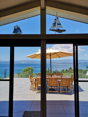 A glass-enclosed patio with a wooden table and chairs, a sailboat model, and a view of the sea and distant land. - Infinity Views - Lake Taupo (free wifi, linen provided) (Omori)