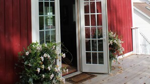 Una granja vermella amb una porta i finestres blanques, un porxo amb felpú i plantes en testos.