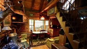 Interior rústico de uma cabana de toras, com área de jantar, escada de madeira e uma grande janela com vista.