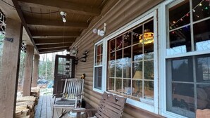 Uma cabana de madeira com varanda, duas cadeiras de balanço, uma mesa pequena e uma janela decorada com luzes de cordão.