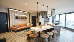 A modern living room with a sofa, coffee table, and wooden stools. There is a dining table with a white top and a vase of fruit. The room has a large window and a view of the city. - B14 Luxury Penthouse down town for groups (Reykjavík)