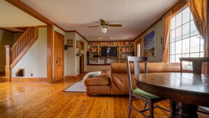 A spacious living room with a wooden staircase, a ceiling fan, a fireplace, a bookshelf, and a dining area with a round table and chairs. - Hyde Park Village Home near STOWE -- 4BR bedroom (Hyde Park)