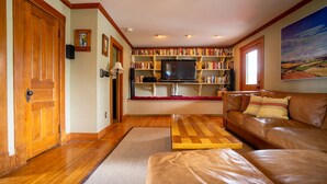 A cozy living room with a leather sofa, a wooden coffee table, a bookshelf filled with books, a flat-screen TV, and a large window. - Hyde Park Village Home near STOWE -- 4BR bedroom (Hyde Park)