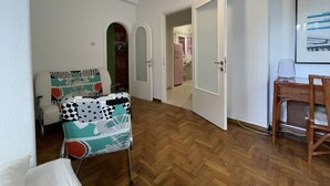 Una habitación con suelo de espiga, un sillón estampado, un escritorio de madera con una silla y una puerta que da a otra habitación.
