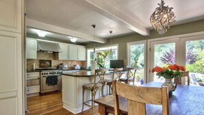 A kitchen with wooden flooring, a dining area with a table and chairs, and a large window with a view of greenery. - CARMEL OASIS-Gorgeous Views 4 Bedroom 2.5- EXCEPTIONAL RATINGS (Carmel-By-The-Sea)