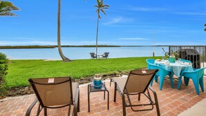 An outdoor patio with a view of the water, featuring a table and chairs, a grill, and a grassy area with palm trees.