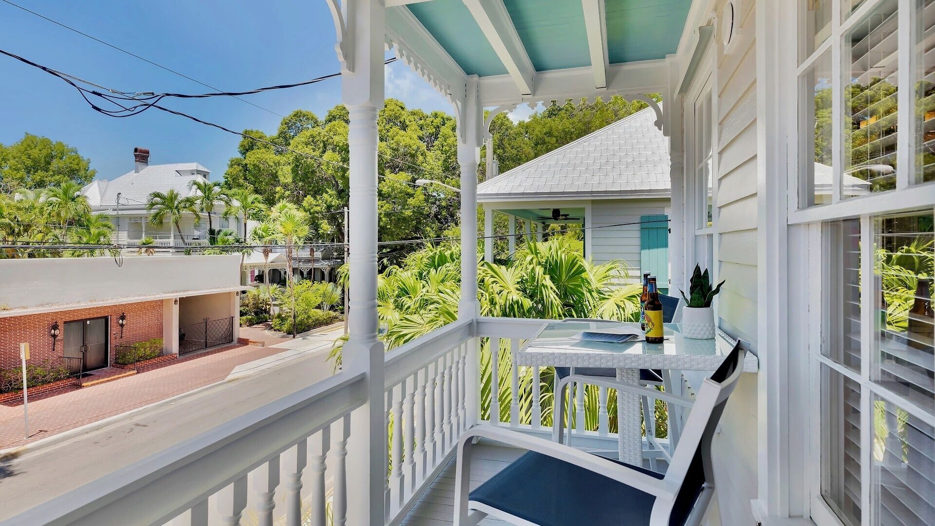 A covered balcony with a table and chairs, surrounded by lush greenery and a view of a residential area.