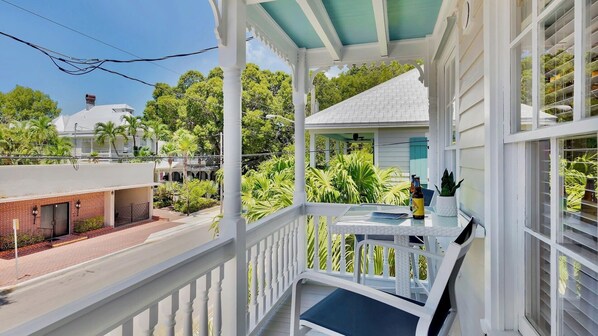 A covered balcony with a table and chairs, surrounded by lush greenery and a view of a residential area.