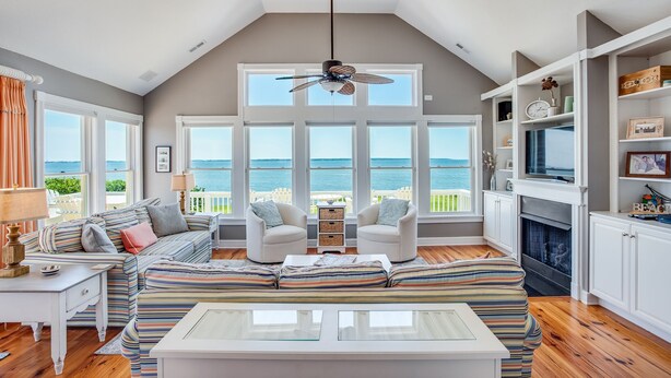 A spacious living room with a large window overlooking the sea, a ceiling fan, and a fireplace. - Tramonti: Soundfront, Heated Pool, Golf (Nags Head)