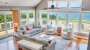 A spacious living room with a large window overlooking a balcony and water. There is a sofa, a coffee table, and two chairs. - Tramonti: Soundfront, Heated Pool, Golf (Nags Head)