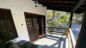 Un porche cubierto con un banco, una silla de mimbre y una puerta que da a un jardín.