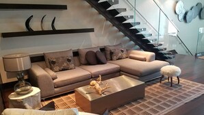 Ein modernes Wohnzimmer mit einem Ecksofa, einem Couchtisch, einer Lampe und einem kleinen Hocker. Im Hintergrund ist eine Treppe mit Glastrennwand zu sehen.