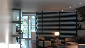 Ein modernes Wohnzimmer mit einer Couch, einem Couchtisch und einer Lampe. Es gibt ein großes Fenster mit Jalousien, eine Tür zu einem anderen Raum und einen Teppich mit Muster auf dem Boden.